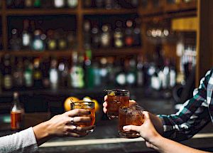 Four people toasting with drinks in a dimly lit bar with shelves of bottles in the background.