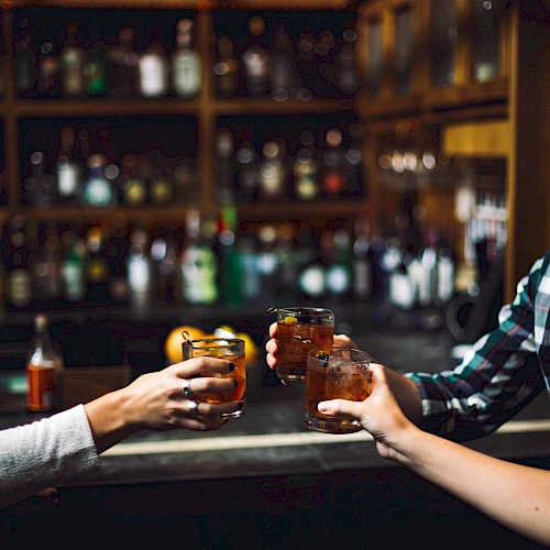 Four people toasting with drinks in a dimly lit bar with shelves of bottles in the background.