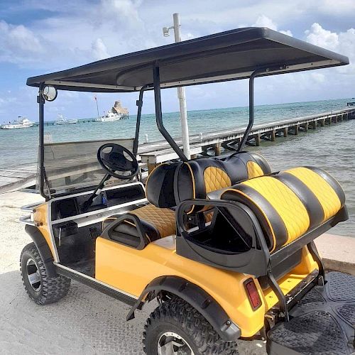 A bright yellow golf cart with black striped seats is parked near a beach with a wooden pier and turquoise water in the background.