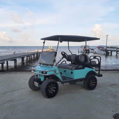 A turquoise golf cart is parked on a sandy beach near a wooden pier, with boats and the ocean visible in the background.