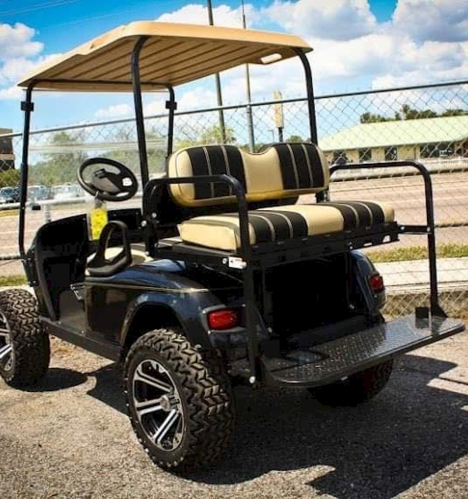 A black golf cart with beige seats and off-road tires is parked on a paved area, next to a chain-link fence.