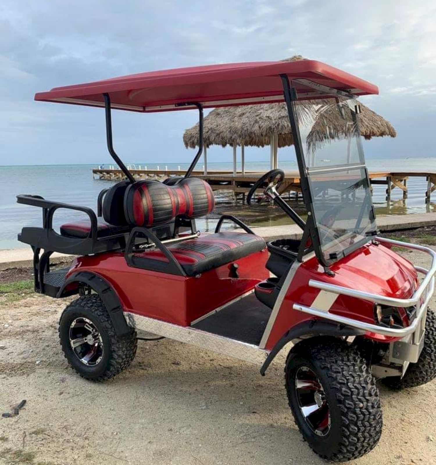 The image features a red and white golf cart with a canopy, parked on a beach near the water, with a wooden pier and thatched-roof structures in the background.