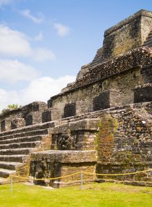 An ancient stone pyramid stands tall under a partly cloudy sky, showcasing its weathered steps and intricate terracing, surrounded by lush greenery.