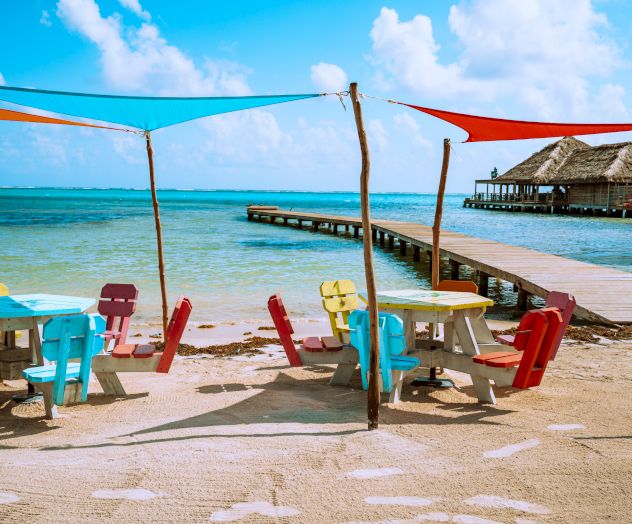 Beach with pastel-colored tables and chairs, shade canopies, and a wooden pier leading to a thatched-roof hut over the water, against a blue sky.