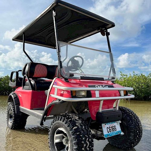 A red off-road golf cart styled vehicle with large tires is parked outdoors. The sky is partly cloudy.