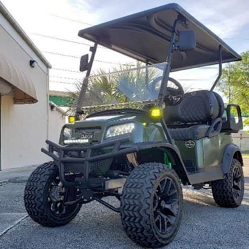 The image shows a green off-road utility vehicle with large tires, a roof, and a seating area, parked near a building on a paved surface.