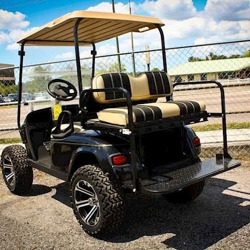 A black golf cart with beige seats and a roof is parked on an asphalt surface next to a chain-link fence, under a partly cloudy sky.