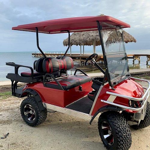 A red and black golf cart is parked on a beach near a pier with thatched-roof structures in the background, under a cloudy sky.