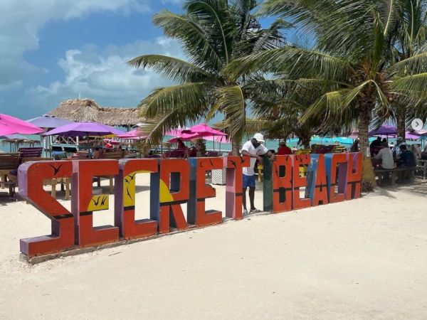 A beach scene with a large "SECRET BEACH" sign, palm trees, colorful umbrellas, and a person standing near the sign.