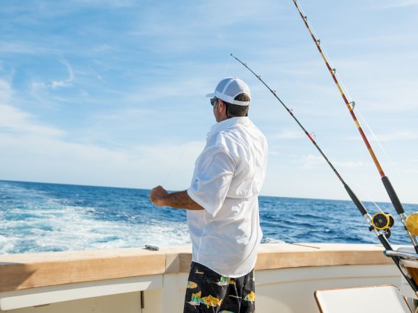 A man in a white shirt and cap is fishing from the back of a boat in the ocean, with fishing rods visible.