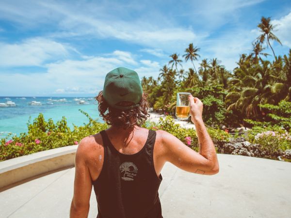 A person in a tank top and cap is holding a drink and enjoying a scenic view of the ocean and palm trees on a sunny day.