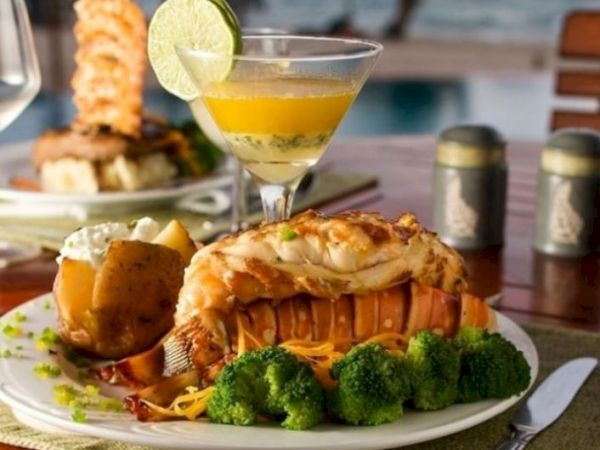 A delicious seafood meal with lobster tail, broccoli, baked potato, and a cocktail garnished with lime, set on a table with an ocean view ending the sentence.