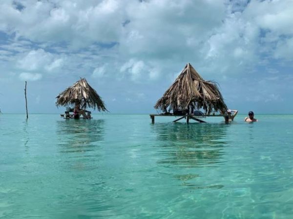 Two thatched-roof huts stand on stilts in clear, shallow turquoise water under a partly cloudy sky; a person is swimming nearby.