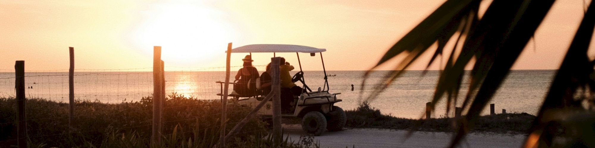 Two people ride a golf cart along a beach at sunset, silhouettes against the glowing horizon.